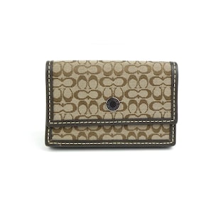 May include: A rectangular wallet featuring a tan and brown repeating logo pattern on a fabric exterior. The wallet has dark brown leather trim with white contrast stitching and a central circular silver-tone snap button closure with Coach branding.