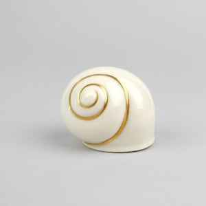May include: A ceramic, off-white seashell-shaped decorative object with a gold spiral design. The glossy finish reflects light, highlighting the elegant curves. The object is a decorative piece.