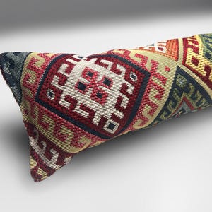 May include: A long, cylindrical pillow with a woven fabric cover. The cover features a repeating geometric pattern in red, cream, navy, yellow, and green. The pillow is likely used for decorative or functional purposes.