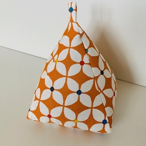 May include: A fabric doorstop in a triangular shape with a small handle. The doorstop has a repeating pattern of white flower shapes on an orange background. Each flower has a coloured dot in the centre, with red, blue, yellow, and black.
