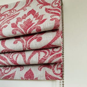 May include: Close-up of a folded window blind featuring a repeating red floral pattern on a light grey background. The blind is made of fabric and has a beaded pull chain on the right. The design is a damask style.