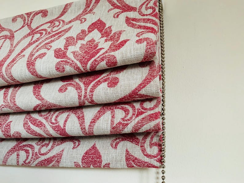 May include: Close-up of a folded window blind featuring a repeating red floral pattern on a light grey background. The blind is made of fabric and has a beaded pull chain on the right. The design is a damask style.