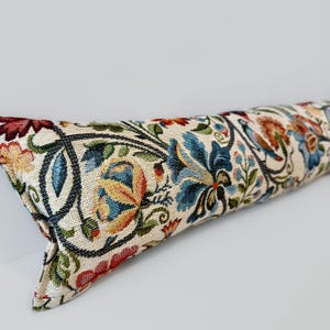 May include: A long, rectangular decorative pillow with a floral pattern. The pillow features a cream-colored background with a repeating design of blue, red, orange, and green flowers and leaves. The pillow is made of a woven fabric.