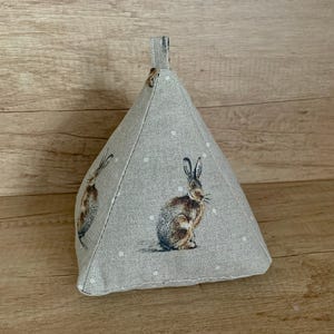 May include: A beige doorstop with a white polka dot pattern and a brown rabbit design. The doorstop is shaped like a pyramid with a loop handle at the top.