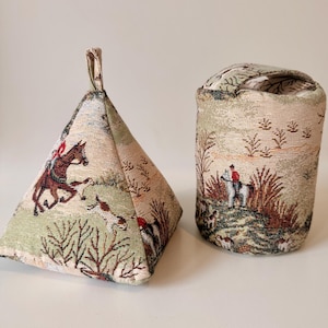 May include: Two fabric doorstops with a hunting scene tapestry design. One is a pyramid shape with a loop handle, the other is cylindrical. The colours are green, brown, and beige, suitable for a country-style home.