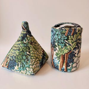 May include: Two fabric doorstops. One is a pyramid shape, the other is cylindrical with a handle. Both are covered in a tapestry-like fabric with a forest scene in shades of green, blue, and brown.
