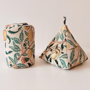 May include: Two decorative fabric items. One is a cylindrical shape, the other is a pyramid shape. Both are beige with a repeating pattern of yellow fruit, green leaves, and brown stems. The pyramid has a small fabric loop at the top.