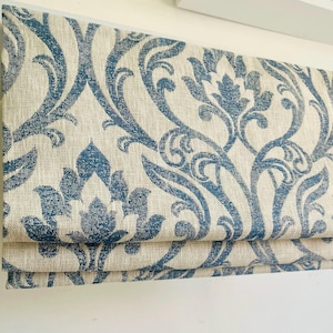 May include: A folded Roman blind with a blue and cream damask design. The fabric has a textured look, with the blue pattern displaying floral and scroll elements. The blind is folded in horizontal layers, indicating a window covering.