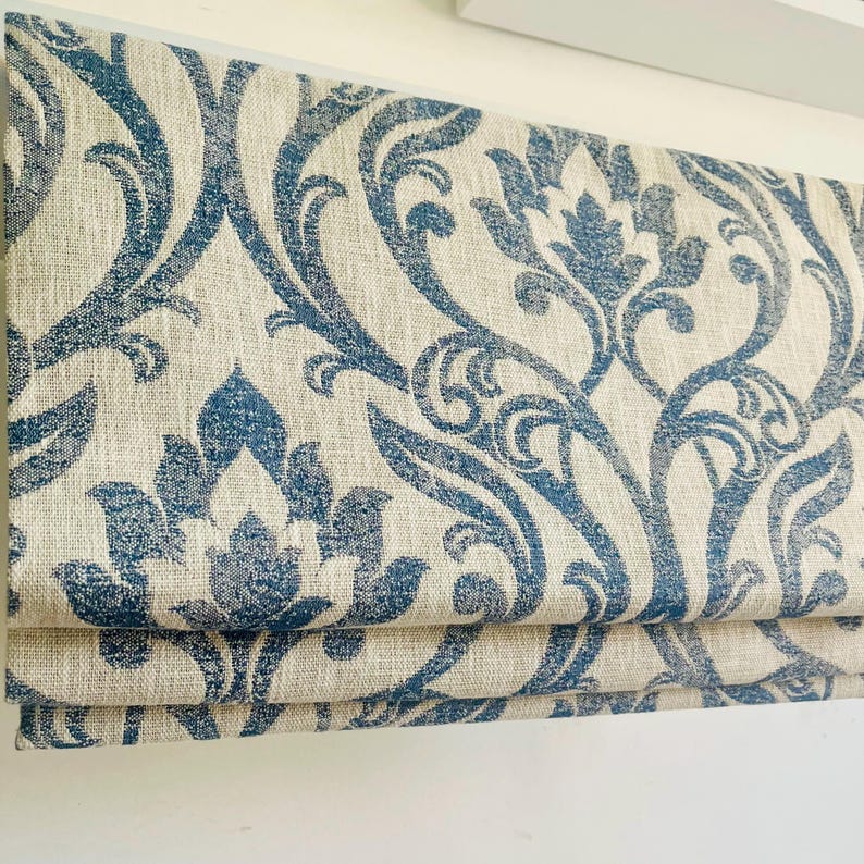May include: A folded Roman blind with a blue and cream damask design. The fabric has a textured look, with the blue pattern displaying floral and scroll elements. The blind is folded in horizontal layers, indicating a window covering.