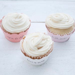 May include: Three white frosted cupcakes with pink wrappers. The wrappers have different text on them: "Megan & Billy", "8.28.2017", and "Love is Sweet".
