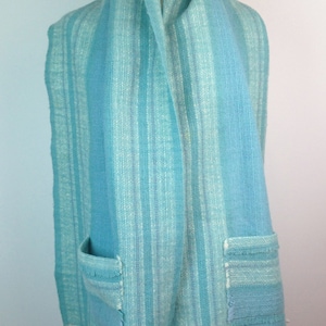 May include: A long, woven scarf in shades of light blue, turquoise, and white, featuring a vertical striped pattern. The scarf includes two patch pockets on the front and is finished with a fringed hem along the bottom edges of the fabric.
