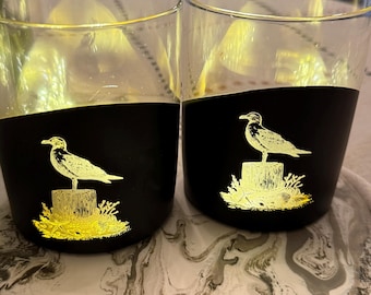 Conjunto de dois copos de uísque gravados com design de gaivota costeira, nas cores preto e dourado.