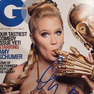 May include: GQ magazine cover featuring Amy Schumer, with the text "OUR TASTIEST COMEDY ISSUE YET!" and a blue signature. Schumer is wearing a brown costume and holding a gold robotic hand. The magazine has a blue "G" logo.