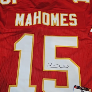 May include: Red football jersey with the name "MAHOMES" in white letters across the chest. The number "15" is prominently displayed in white with gold trim. An autograph is visible on the jersey, along with a Global Authentics certificate.