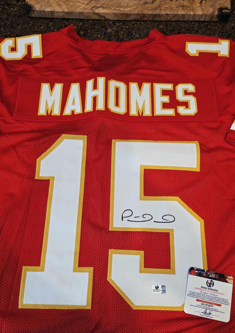 May include: Red football jersey with the name "MAHOMES" in white letters across the chest. The number "15" is prominently displayed in white with gold trim. An autograph is visible on the jersey, along with a Global Authentics certificate.