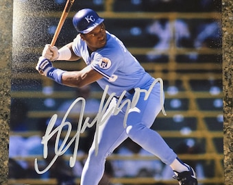 May include: A glossy photograph of a baseball player in a light blue uniform with a KC cap, holding a wooden bat in a batting stance. The image features a large silver signature across the centre and a circular holographic sticker in the corner.