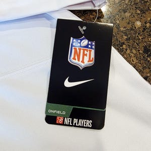May include: White garment with a red stripe and a black tag. The tag features the NFL logo, a Nike swoosh, and the text "ONFIELD NFL PLAYERS". The garment appears to be athletic wear.