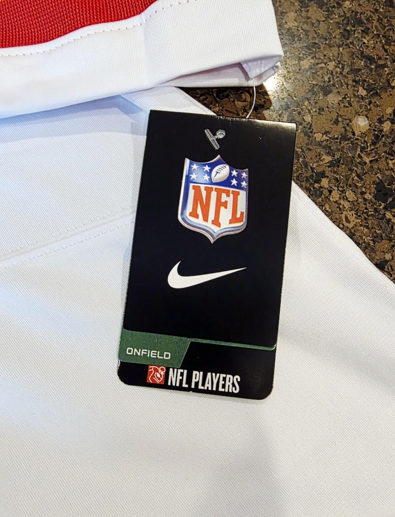 May include: White garment with a red stripe and a black tag. The tag features the NFL logo, a Nike swoosh, and the text "ONFIELD NFL PLAYERS". The garment appears to be athletic wear.
