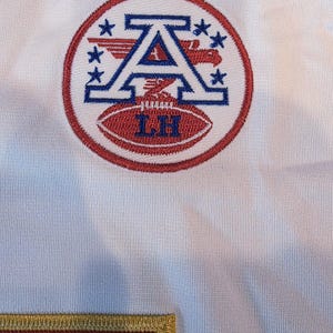May include: White jersey with a circular emblem featuring a blue "A" with wings and stars, and a red football with "LH" embroidered on it. The emblem is surrounded by a red circle. A maroon patch with gold trim is visible.