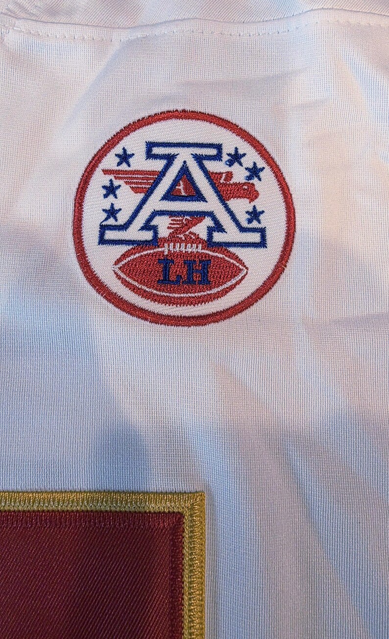 May include: White jersey with a circular emblem featuring a blue "A" with wings and stars, and a red football with "LH" embroidered on it. The emblem is surrounded by a red circle. A maroon patch with gold trim is visible.