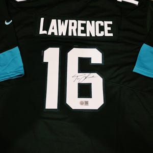 May include: A black sports jersey featuring the name LAWRENCE and the number 16 in white lettering on the back. The jersey includes teal accents on the sleeves and collar, a small Nike logo, and a black ink autograph on the number 16.