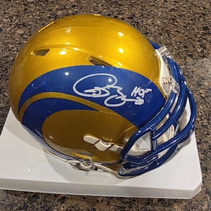 Puede incluir: Un casco de fútbol americano dorado y azul con una firma y el texto "HOT 20". El casco tiene una máscara azul y está sobre una superficie blanca. El casco tiene un acabado brillante.