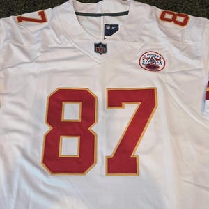 May include: White football jersey with red and gold accents. Features the number 87 in large red numerals with gold trim on the front. The number 8 and 7 are also on the shoulders. Includes an NFL logo and a team logo.