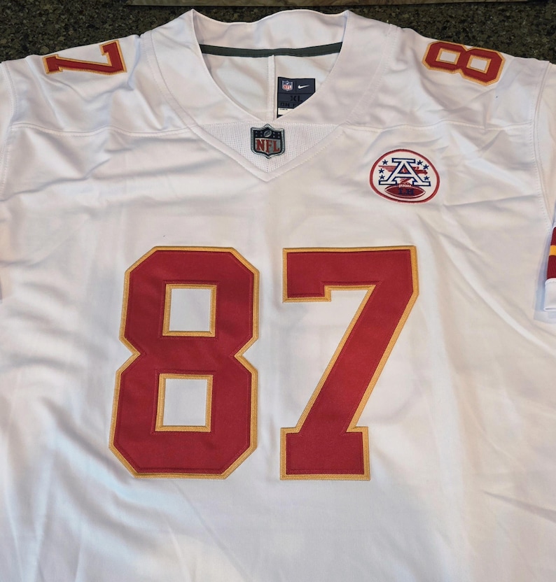 May include: White football jersey with red and gold accents. Features the number 87 in large red numerals with gold trim on the front. The number 8 and 7 are also on the shoulders. Includes an NFL logo and a team logo.