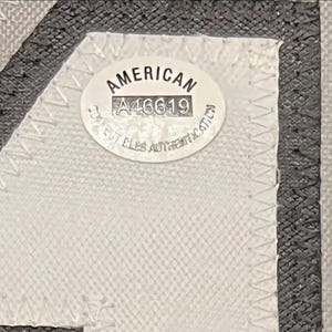 May include: Close-up of a black and gray jersey with white stitching and a white patch. The patch reads "AMERICAN" and includes the number "A46619" and text that is difficult to read.