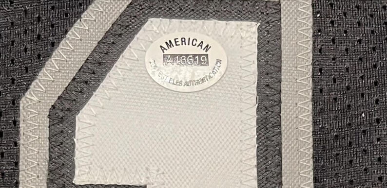 May include: Close-up of a black and gray jersey with white stitching and a white patch. The patch reads "AMERICAN" and includes the number "A46619" and text that is difficult to read.
