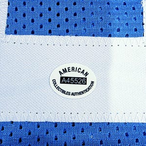 May include: Close-up of a blue mesh fabric with a white stripe and stitching. An oval sticker with the text "AMERICAN COLLECTIBLES AUTHENTICATION" and the number "A45526" is visible. The fabric has small, evenly spaced holes.