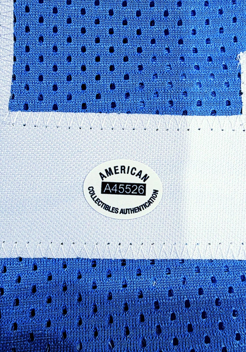 May include: Close-up of a blue mesh fabric with a white stripe and stitching. An oval sticker with the text "AMERICAN COLLECTIBLES AUTHENTICATION" and the number "A45526" is visible. The fabric has small, evenly spaced holes.