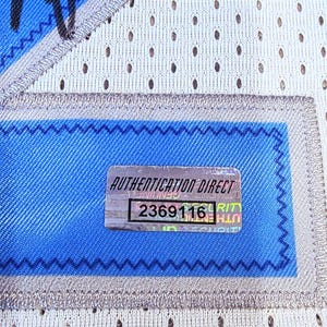 May include: Close-up of a section of a white mesh jersey with blue accents. A rectangular patch with the text "AUTHENTICATION DIRECT" and the number "2369116" is visible. The jersey has a blue and silver trim.