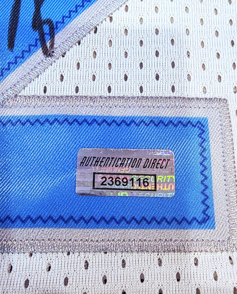 May include: Close-up of a section of a white mesh jersey with blue accents. A rectangular patch with the text "AUTHENTICATION DIRECT" and the number "2369116" is visible. The jersey has a blue and silver trim.