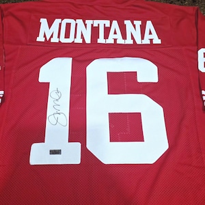 May include: A red sports jersey featuring the name MONTANA in white block letters across the upper back. The number 16 is printed in large white digits below, with a black ink signature on the number 1 and a small authentication sticker attached.