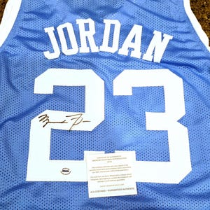 May include: Light blue mesh jersey with the name "JORDAN" and the number "23" in white. The jersey has a white collar and armbands. Includes a signature and an authentication certificate.