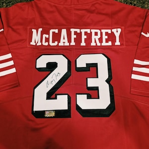May include: A red sports jersey featuring the name McCaffrey and the number 23 in white with black outlines on the back. A black ink signature is visible on the number 2, and a small holographic authentication sticker is attached to the number 3.