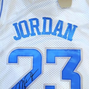May include: White mesh jersey with the name "JORDAN" in light blue block letters across the chest. The number "23" is prominently displayed in light blue with a silver outline. A signature is visible on the number 23. A tag with the Jumpman logo hangs from the jersey.