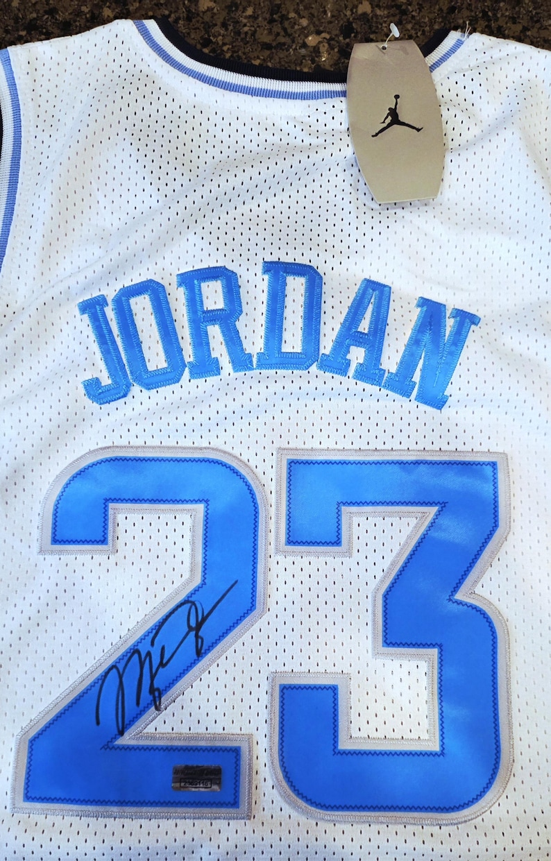 May include: White mesh jersey with the name "JORDAN" in light blue block letters across the chest. The number "23" is prominently displayed in light blue with a silver outline. A signature is visible on the number 23. A tag with the Jumpman logo hangs from the jersey.