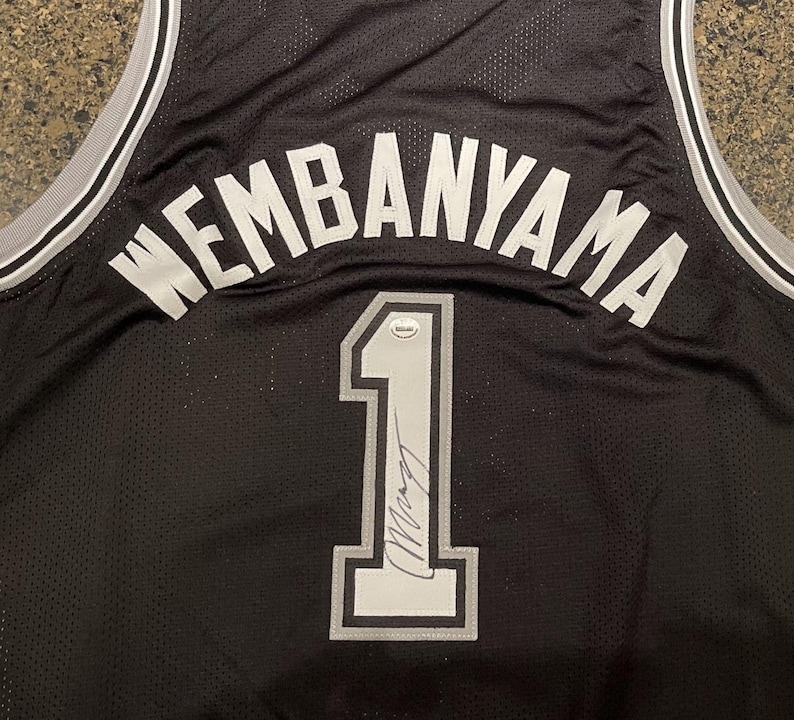 May include: Black basketball jersey with the name "WEMBANYAMA" in white block letters across the back. The number "1" is displayed in white with a gray outline, and a signature is visible below the number.