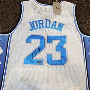 May include: White mesh basketball jersey with the name "JORDAN" in light blue across the chest and the number "23" in light blue. The jersey has a signature below the number and a tag with a black Jumpman logo.