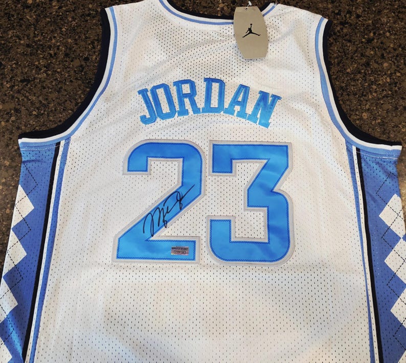May include: White mesh basketball jersey with the name "JORDAN" in light blue across the chest and the number "23" in light blue. The jersey has a signature below the number and a tag with a black Jumpman logo.