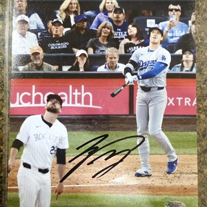 Puede incluir: Fotografía firmada de un jugador de béisbol con el uniforme de los Dodgers de Los Ángeles, en pleno swing, con un bateador de los Rockies de Colorado en primer plano. El fondo muestra espectadores en las gradas. La fotografía está en una funda protectora.