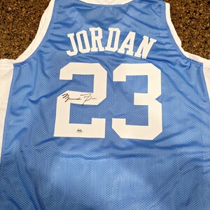 May include: Light blue basketball jersey with the name "JORDAN" and the number "23" in white. The jersey has a white trim and a signature on the number 23. The jersey is made of a breathable mesh material.