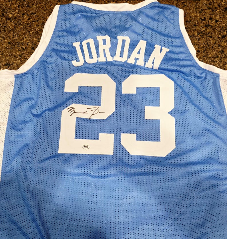 May include: Light blue basketball jersey with the name "JORDAN" and the number "23" in white. The jersey has a white trim and a signature on the number 23. The jersey is made of a breathable mesh material.