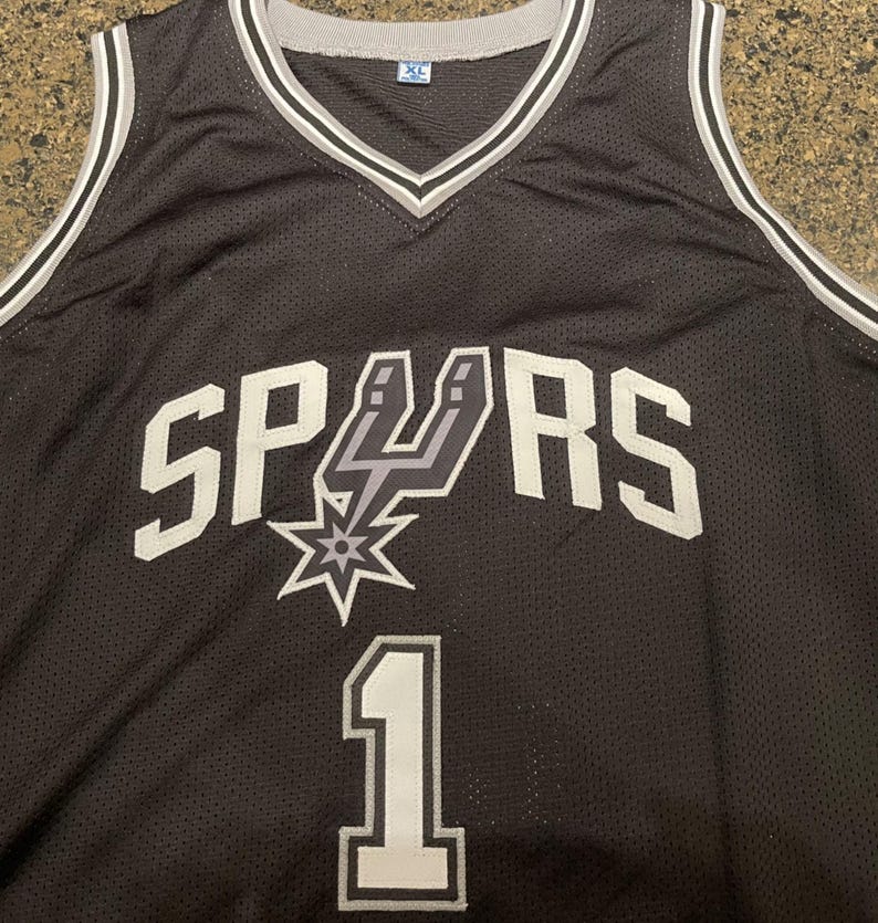 May include: Black XL basketball jersey with the word "SPURS" in white block letters. The jersey features the Spurs logo and the number "1" in white. The jersey has a V-neck and white and gray trim.