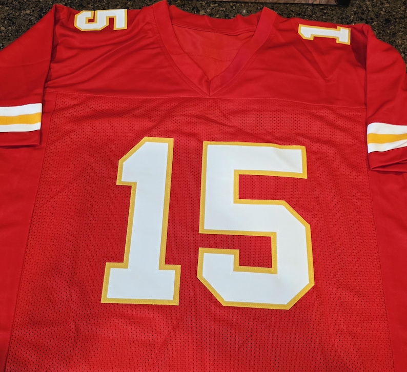 May include: Red football jersey with white and yellow accents. The jersey features the number 15 in large white block letters with a yellow outline on the front. The sleeves have white and yellow stripes.