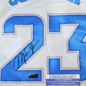 May include: White mesh jersey with the name "JORDAN" in blue lettering across the top. The number "23" is prominently displayed in blue with a gray outline. An autograph is visible on the jersey. A "AUTHENTICATION DIRECT" certificate is attached.