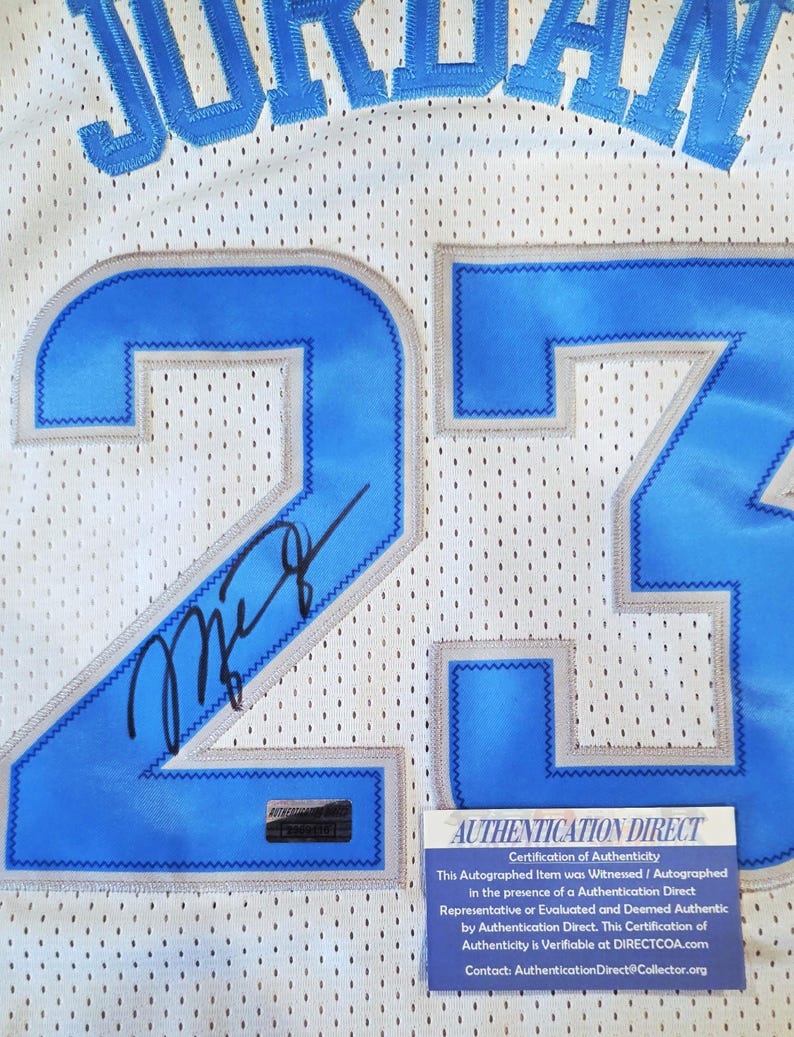 May include: White mesh jersey with the name "JORDAN" in blue lettering across the top. The number "23" is prominently displayed in blue with a gray outline. An autograph is visible on the jersey. A "AUTHENTICATION DIRECT" certificate is attached.