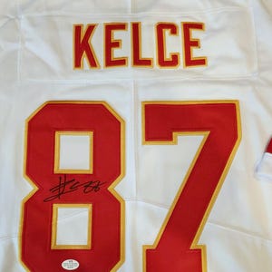 May include: White football jersey with the name "KELCE" in red and gold lettering. The number "87" is prominently displayed in red with gold trim. Includes an autograph and a certification sticker.